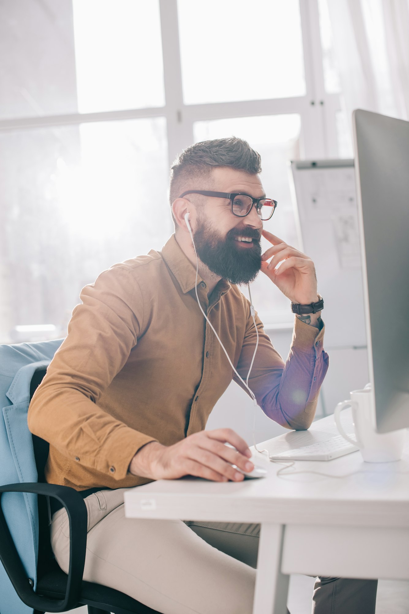 smiling bearded adult businessman in earphones sitting and working at computer desk in office